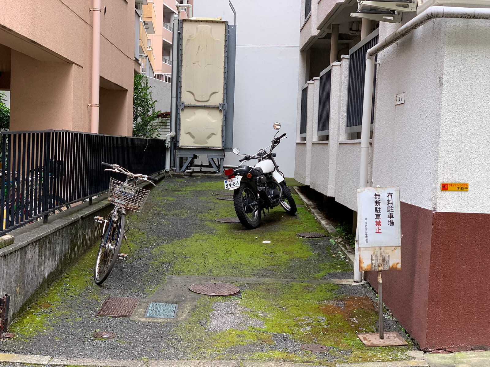 A Kawasaki trail bike and a city bicycle in a moss-covered alley between apartment buildings, with a no-parking sign