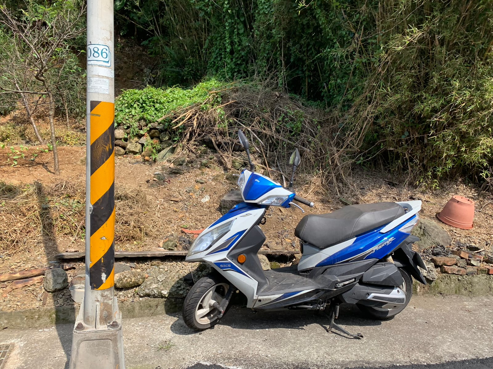 A blue and white SYM Fighter scooter parked below a hillside of bamboo, beside a yellow-and-black striped pole