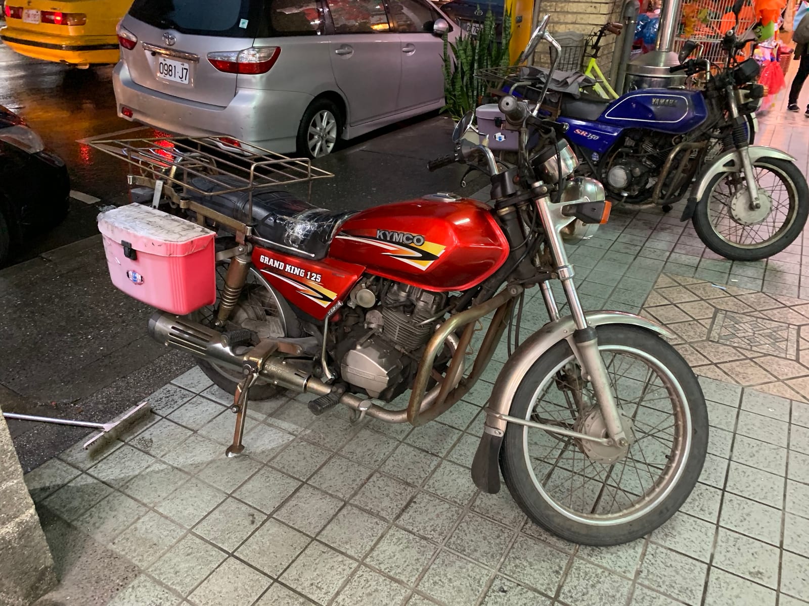 A red KYMCO Grand King 125 with a pink plastic delivery box and a chrome rear rack, parked on a wet pavement at night