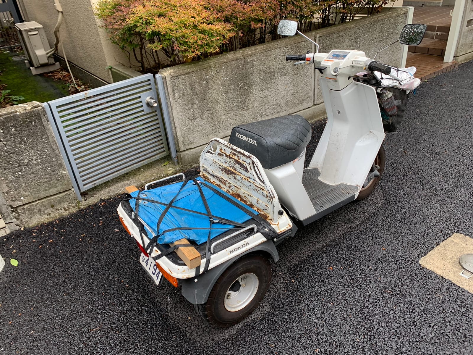 A white Honda three-wheeled delivery scooter with a blue tarp strapped to its rear deck, parked beside a garden wall