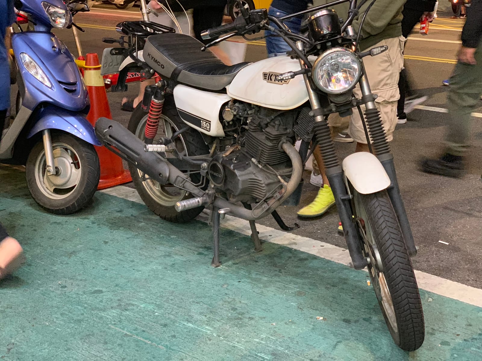 A white KYMCO 150 with a round headlamp and red-sprung shocks, parked on a green painted kerb beside a traffic cone at night