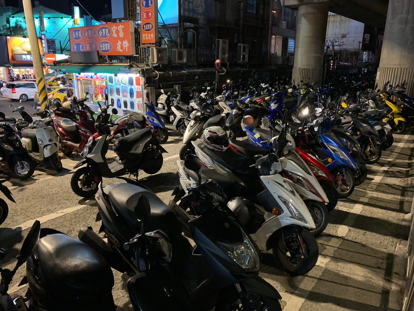 Dozens of scooters packed nose to tail in a lot under an elevated expressway at night, a luggage shop lit up behind them