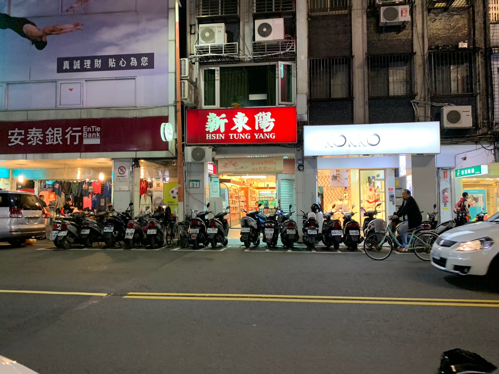 A row of scooters lined up outside shopfronts at night, under a red Hsin Tung Yang sign and a bank
