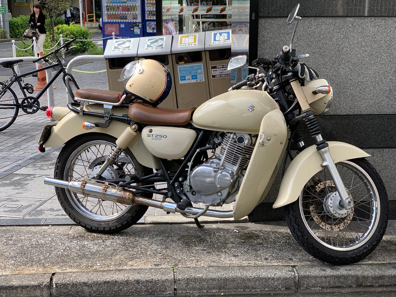 A cream Suzuki ST250 with a brown seat and a matching open-face helmet, parked outside a convenience store