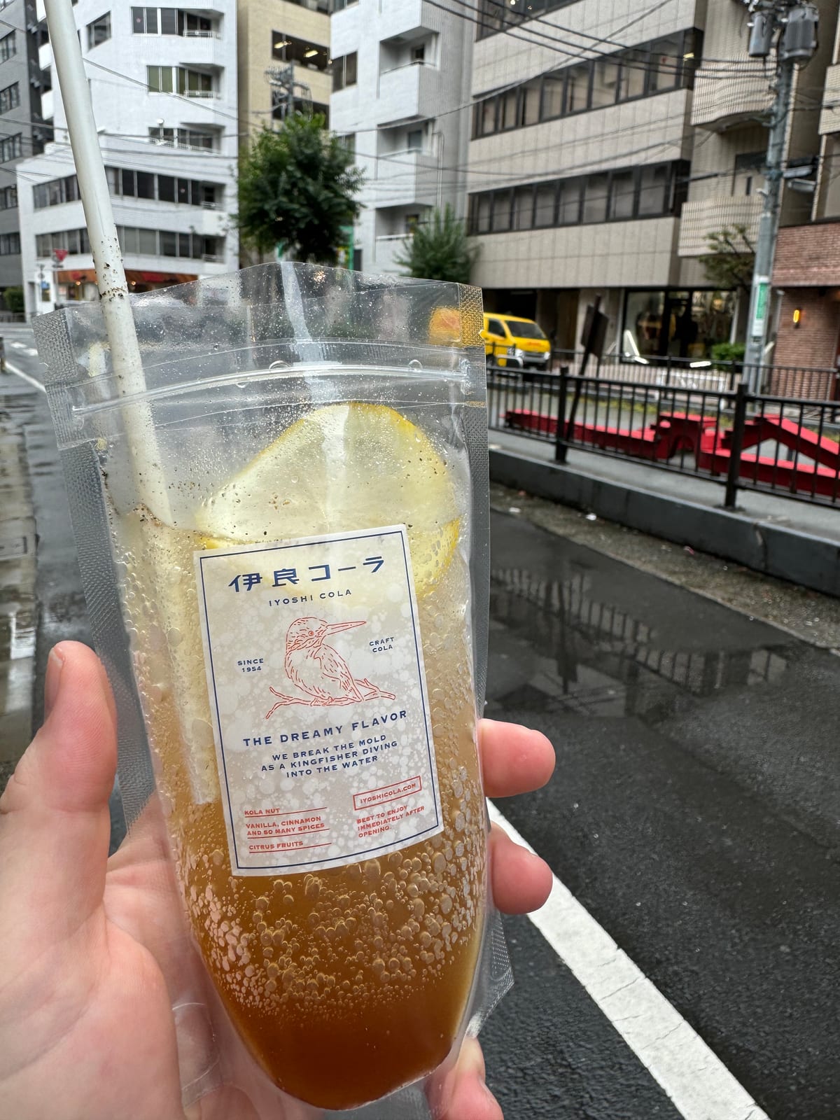 A clear pouch of Iyoshi Cola with a lemon slice and paper straw, held up over a wet Tokyo street