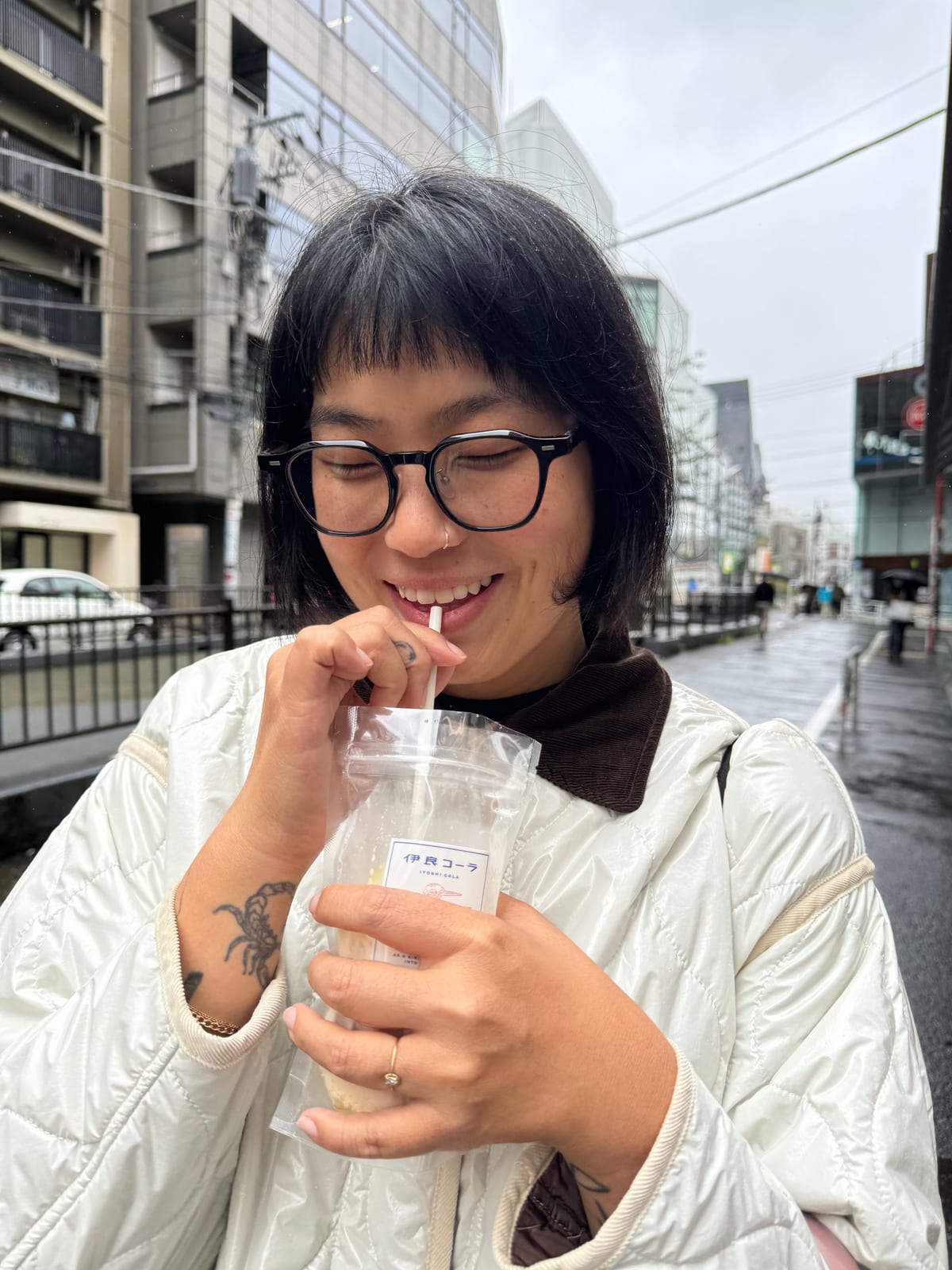 A customer in glasses and a cream quilted jacket drinking Iyoshi Cola through a paper straw, eyes closed, smiling