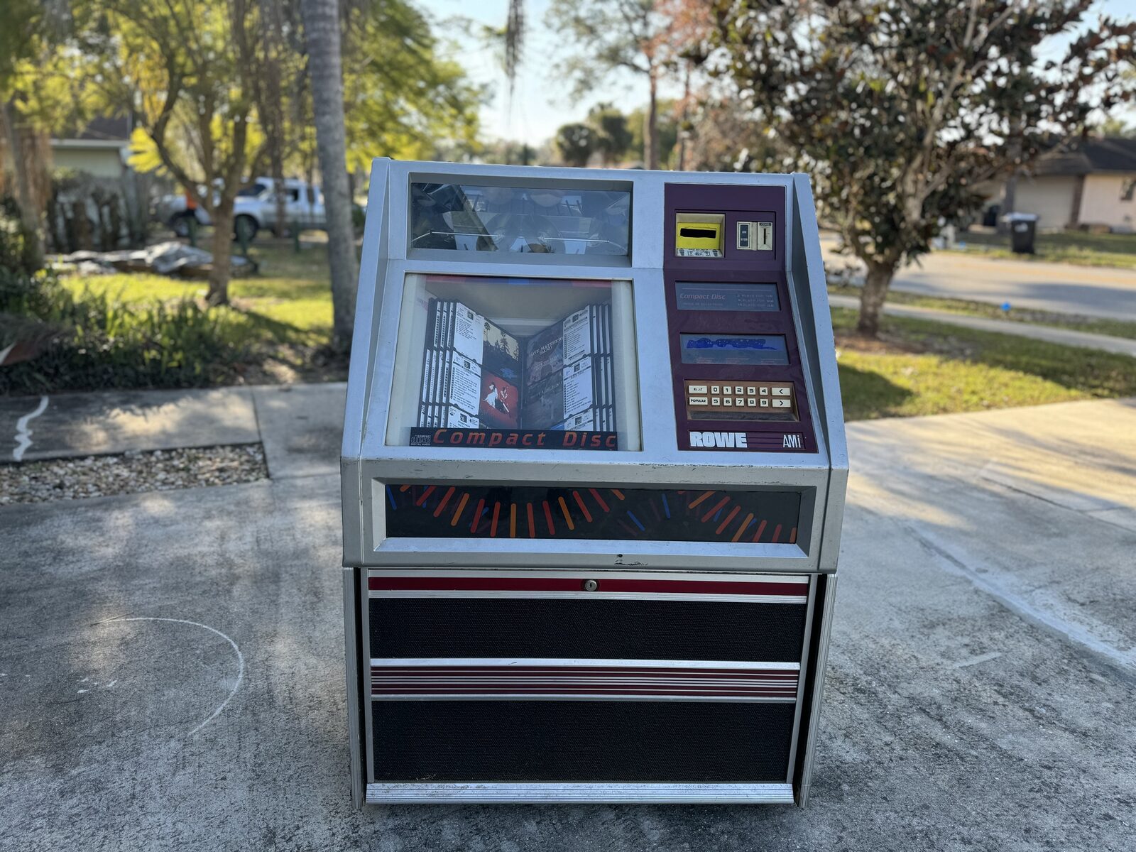 The full Rowe AMI jukebox on a driveway: a CD carousel window on top, the Compact Disc title rack, a maroon keypad panel, and black speaker grilles below